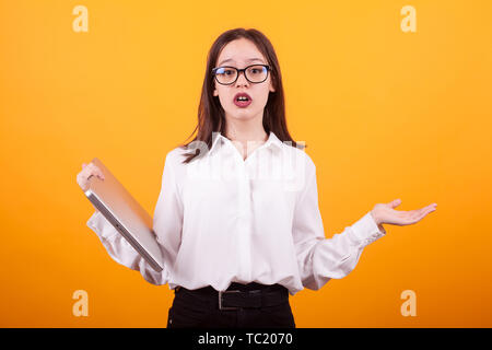 Ritratto di carino ragazza giovane con occhiali cercando cercando confuso e azienda laptop in studio su sfondo giallo. Yong ragazza utilizzando la tecnologia moderna. Foto Stock