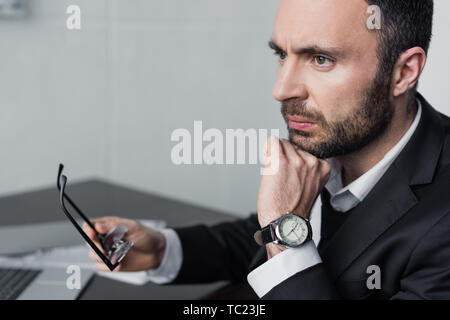 Sconvolto barbuto imprenditore seduti al posto di lavoro, tenendo gli occhiali e guardando lontano Foto Stock