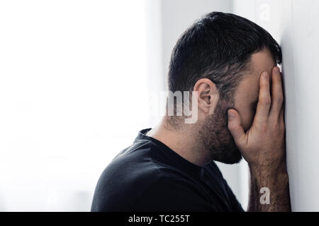 Triste adulto uomo in piedi da muro bianco e che copre il viso con le mani Foto Stock