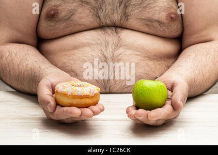 Uomo obesi tiene in mano una mela e un concetto di ciambella di una dura scelta tra alternative opposte (cibo sano vs cibo dolce) Foto Stock