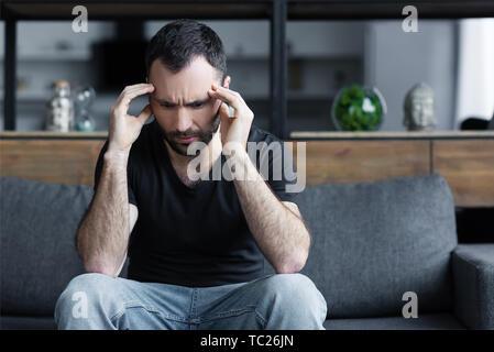 Triste uomo adulto foratura mani vicino a capo mentre è seduto sul divano grigio a casa Foto Stock