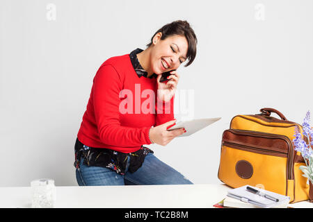 Vista frontale della bella donna con coda di cavallo seduti su uno sgabello mentre si utilizza il telefono e tenendo un tablet pc contro uno sfondo bianco Foto Stock