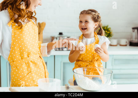 Eccitato figlia emotiva tenendo le uova mentre madre in giallo polka dot grembiule puntando con il dito in cucina Foto Stock