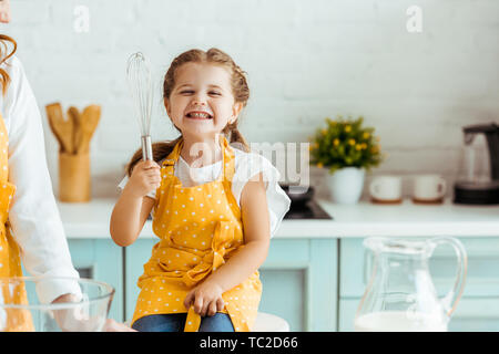 Eccitata la figlia a pois grembiule giallo tenendo palloncino frusta accanto alla madre in cucina Foto Stock