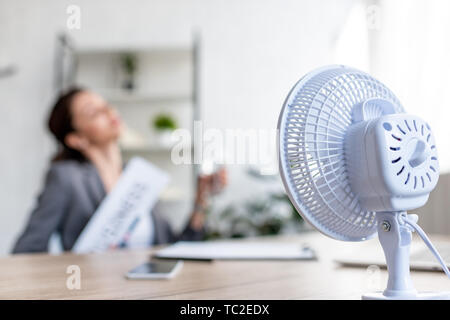 Messa a fuoco selettiva di imprenditrice che soffrono di calore mentre seduto vicino a ventola elettrica Foto Stock