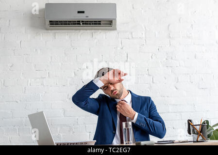 Bello imprenditore soffre di calore mentre seduti al posto di lavoro sotto il condizionatore d'aria Foto Stock