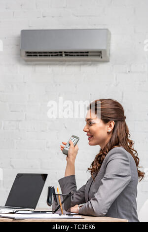 Sorridente imprenditrice holding di controllo remoto mentre seduto sotto il condizionatore d'aria al lavoro Foto Stock