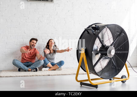 Emozionato l uomo e la donna seduta sul pavimento con le mani tese nella parte anteriore del ventilatore elettrico Foto Stock