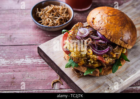 Fritti e croccanti insetto mealworm burger su un panino fresco servito con insalata di passamaneria e un piatto di lato di worms su una tavola in legno rustico con spazio di copia Foto Stock