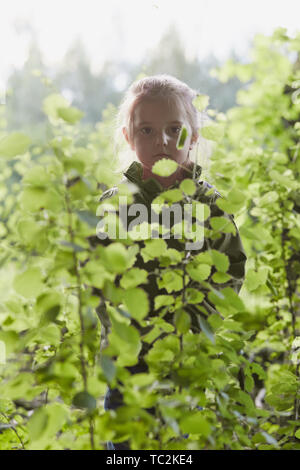 Bambina in piedi nel bosco dietro le foglie, guardando la telecamera, indossando vestiti verde. Candide persone, veri momenti e situazioni reali Foto Stock