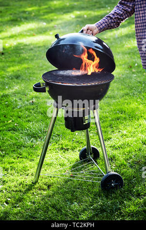 L'uomo il sollevamento del coperchio di un barbecue portatile incendio in un bollitore grill per verificare se i carboni sono pronti per iniziare la cottura del cibo all'aperto su erba verde Foto Stock