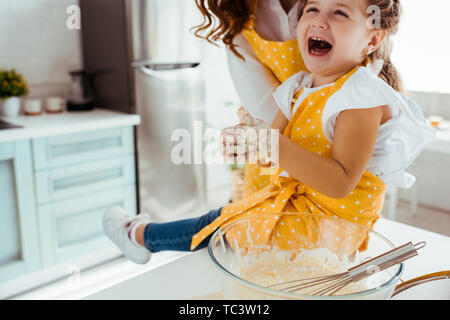 Bambino eccitato in polka dot grembiule con la pasta su mani di ridere mentre è seduto accanto alla madre sul tavolo da cucina Foto Stock