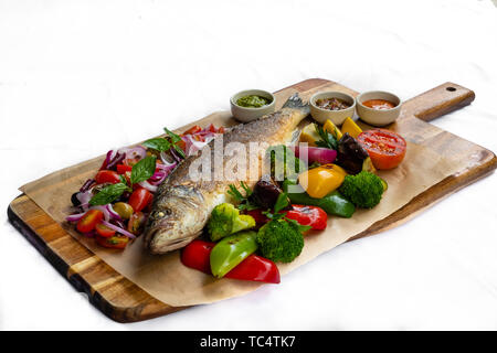 Close up di grigliate di mare mediterraneo bass pesce intero isolato scheda di legno naturale luce posteriore verdure arrosto patate al forno mescolare i pomodori ciliegia basilico Foto Stock