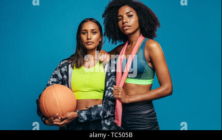 Due giovani femmine nell'abbigliamento sportivo in piedi insieme con la pallacanestro e banda di resistenza. Razza mista di donne in costume fitness guardando la fotocamera con confi Foto Stock