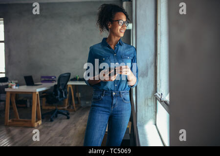 Donna sorridente in casuals in piedi in ufficio e guardando fuori dalla finestra. Imprenditrice con il cellulare in mano guardando fuori dalla finestra. Foto Stock