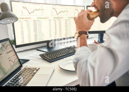 Godere il pranzo. Close-up del barbuto trader è mangiare sandwich freschi e guardando lo schermo del monitor con i dati finanziari mentre è seduto nel suo ufficio moderno. Pausa caffè. Concetto di cibo. Il concetto di business. Concetto di commercio Foto Stock