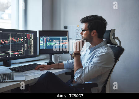 Intensa giornata di lavoro. Vista laterale di successo del commerciante o imprenditore nel formale e indossare occhiali a lavorare con grafici e report di mercato su schermi di computer nel suo ufficio moderno. Stock broker. Mercato Forex. Concetto di commercio Foto Stock