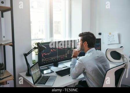 Trading sui mercati di tutto il mondo. Giovani stock broker del mercato analizzando i dati e grafici su più schermi di computer mentre è seduto in un ufficio moderno. Stock Exchange. Concetto di commercio. Concetto di investimento Foto Stock