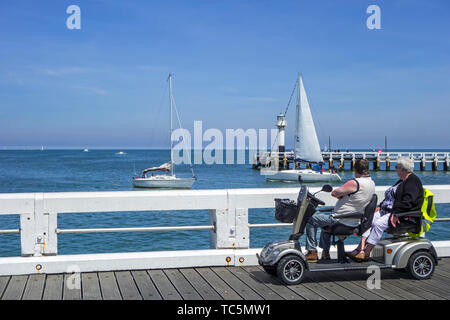 Disattivato i pensionati in duo due persona scooter di mobilità a guardare le barche a vela in mare dal molo al centro balneare in estate Foto Stock