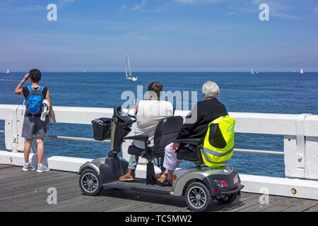 Disattivato i pensionati in duo due persona scooter di mobilità a guardare le barche a vela in mare dal molo al centro balneare in estate Foto Stock