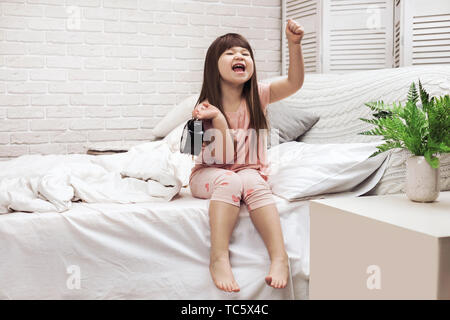 Sorridente bambina ragazza in pigiama con orologio a letto. buona mattina Foto Stock