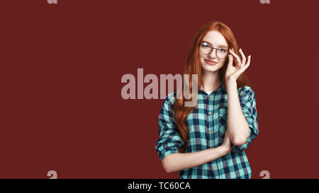 Close up della cute redhead ragazza con i capelli lunghi toccando specs e guardando la telecamera indossando camicia a scacchi isolate su sfondo rosso. Inquadratura orizzontale Foto Stock