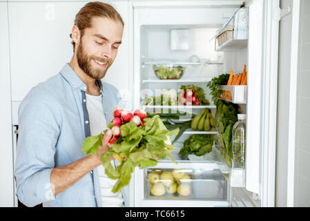 Giovane uomo vegano la scelta di cosa cucinare, tenendo le verdure fresche dal frigorifero a casa Foto Stock