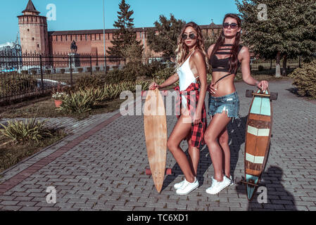 Due ragazze fidanzate, estate città skateboard, longboard costume da bagno shorts. Ponendo il resto comunicare i capelli lunghi conciate di figura. Gli alberi di sfondo edificio Foto Stock