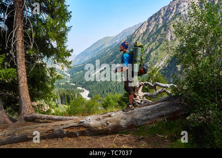 Barbuto fotografo maschio con uno zaino sulla schiena e una fotocamera reflex nelle sue mani. Uomo in una maglietta blu sorge sul bordo di un sentiero di montagna. Guy Foto Stock
