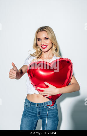 Allegro ragazza bionda holding rosso a forma di cuore mentre il palloncino mostra pollice in alto su bianco Foto Stock