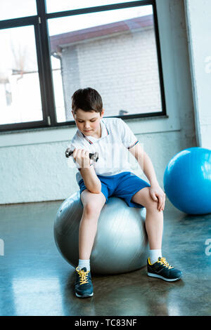 Carino Ragazzo seduto sulla sfera di fitness e lavorare fuori con il manubrio in palestra Foto Stock