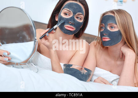 Giovani donne che indossano gli asciugamani resto insieme a casa di cura di bellezza giacente sul letto guardando lo specchio nero di applicare la maschera di argilla sulla faccia concentrata di close-up Foto Stock