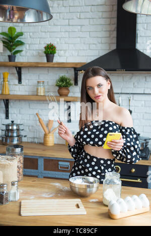 Una giovane e bella bruna ragazza in un abito a pois massaggia un impasto e dischi in uova per una torta. Donna cuochi in cucina secondo la ricetta. Foto Stock