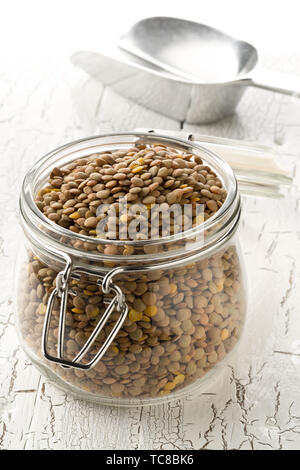 Raw, secco, non cotte lenticchia marrone legumi in vetro vaso di storage su legno bianco sullo sfondo della tabella con il fuoco selettivo Foto Stock