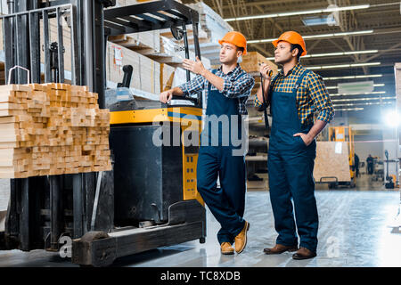 Bello il magazzino multiculturale dei lavoratori nei caschi in piedi vicino al carrello macchina in magazzino Foto Stock