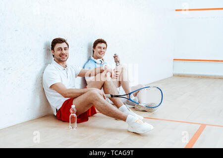 Due sorridente squash giocatori seduti sul pavimento in quattro-walled corte Foto Stock