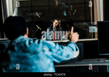 Messa a fuoco selettiva di razza mista produttore del suono che mostra il pollice fino a donna cantare in studio di registrazione Foto Stock