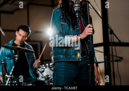 Messa a fuoco selettiva di donna cantare in studio di registrazione mentre razza mista musicista suonando la batteria Foto Stock