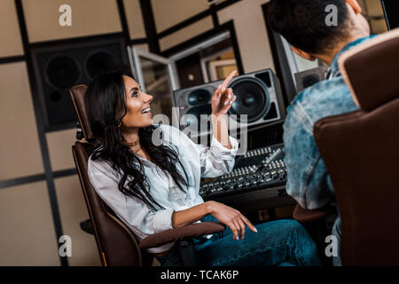 Piuttosto sorridente giovane produttore puntando con il dito nei pressi di razza mista collega in studio di registrazione Foto Stock