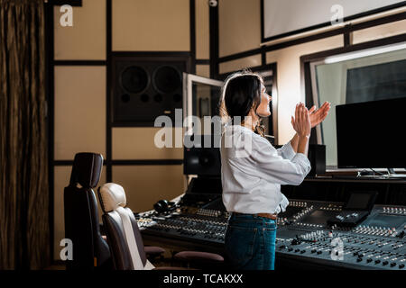 Attraente produttore audio applaudire in studio di registrazione nei pressi di console di mixaggio Foto Stock