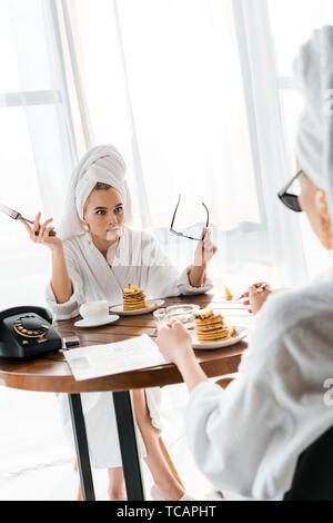 Scioccato donna elegante in accappatoio e di gioielli con asciugamano sulla testa forcella di supporto e gli occhiali da sole e fissando amico durante la prima colazione Foto Stock