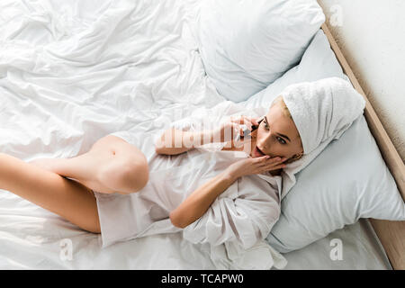 Vista superiore della donna scioccato in gioielleria con asciugamano sulla testa giacente in letto bianco e parlando sullo smartphone Foto Stock