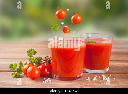 I succhi di pomodoro con prezzemolo in bicchieri e pomodori ciliegia di cadere in vetro sul tavolo di legno. Foto Stock