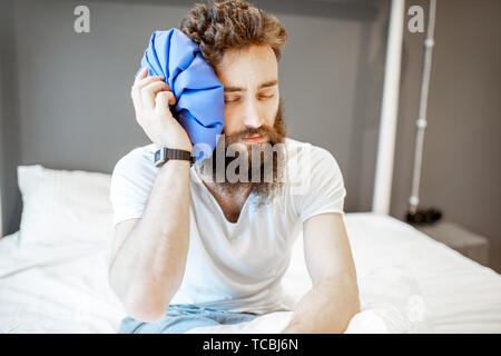 L'uomo sensazione di cattivo, seduta sul letto con mal di testa e applicando il sacchetto del ghiaccio Foto Stock