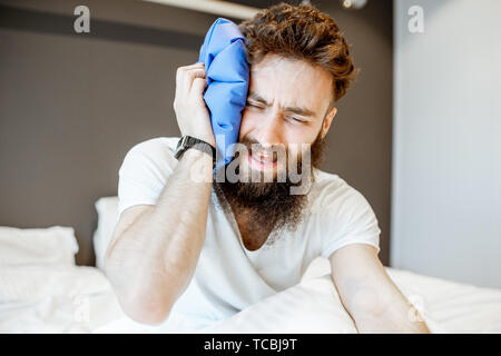 L'uomo sensazione di cattivo, seduta sul letto con mal di testa e applicando il sacchetto del ghiaccio Foto Stock