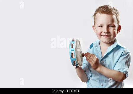 Allegro ragazzo in una maglietta blu tenendo un tamburello e sorridente su uno sfondo luminoso Foto Stock