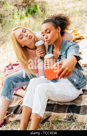 Due gioiosa amici multiculturale con bicchieri di carta di caffè tenendo selfie nella foresta Foto Stock