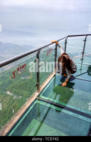 Giovani caucasici donna bianco inginocchiato sul vetro di altissima Sky camminare in montagna Tianmen, vista sulla valle e sulle montagne verso il basso al di sotto, Zhangjiajie, Hunan, Foto Stock