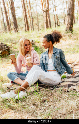 Due amici multietnica seduto su plaid Coperte e tenendo la carta tazze di caffè Foto Stock