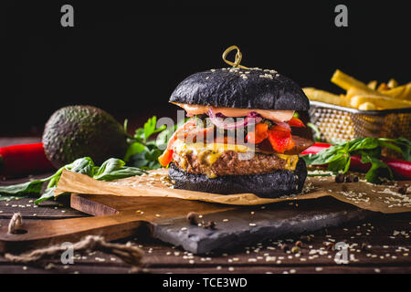 Close-up di gustosi hamburger di manzo con black bun, salsiccia, grigliate di peperone, Chimichurri salsa marinata e cipolla rossa e le patatine fritte in un taglio di legno Foto Stock